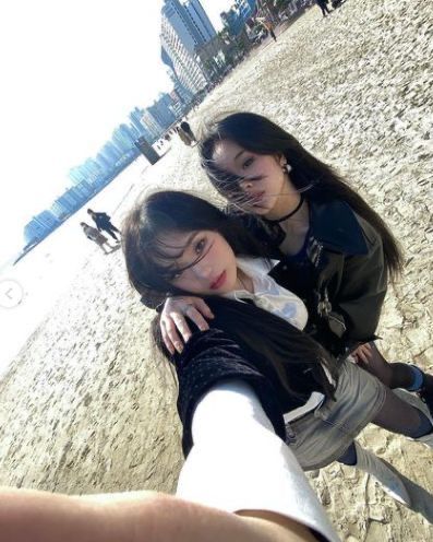 a selfie taken by two young women on a beach with a city skyline in the background. they are both wearing black and white outfits. the photo is taken from a low angle, making the beach and city appear vast. one woman has her arm around the other as they both smile at the camera.