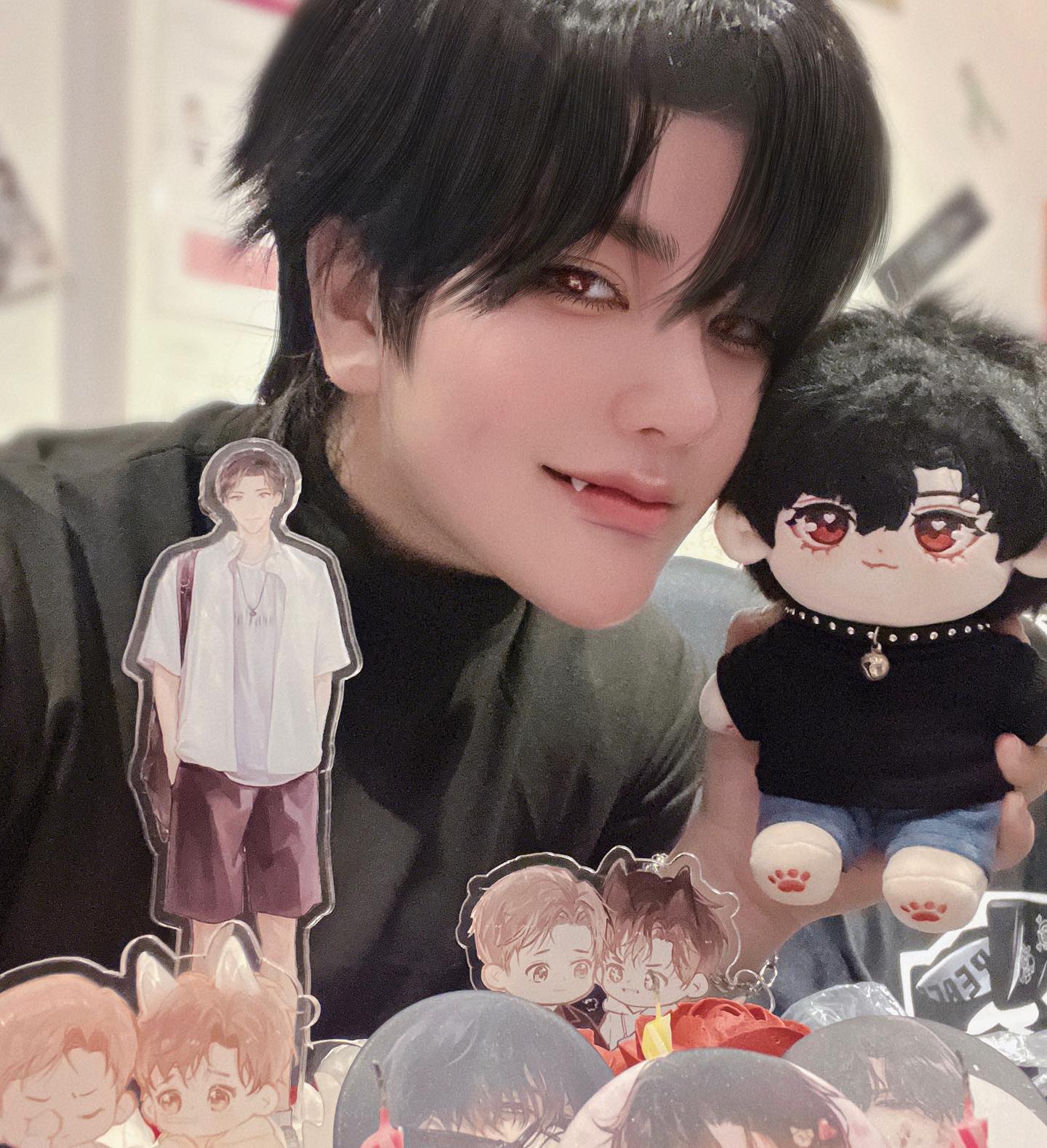 a person cosplaying a vampire holds a plush doll of an anime character. they are surrounded by several figures of different anime characters. the plush doll has black hair and red eyes, and is wearing a black shirt, jeans, and a silver chain. the person is also wearing a black shirt and has red eyes. the background is blurry, and the focus is on the person and the plush doll.