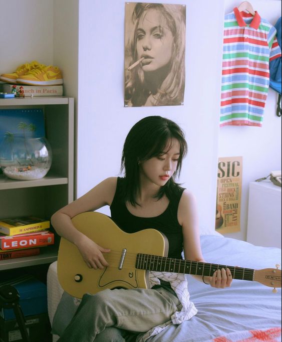 a young woman with black hair and a black tank top sits on a bed playing a yellow guitar. she is surrounded by music paraphernalia, including a bookshelf with books and magazines. she is in a welllit bedroom with a poster of a woman and a striped polo shirt hanging on the wall. the image captures the simple joy and beauty of making music.