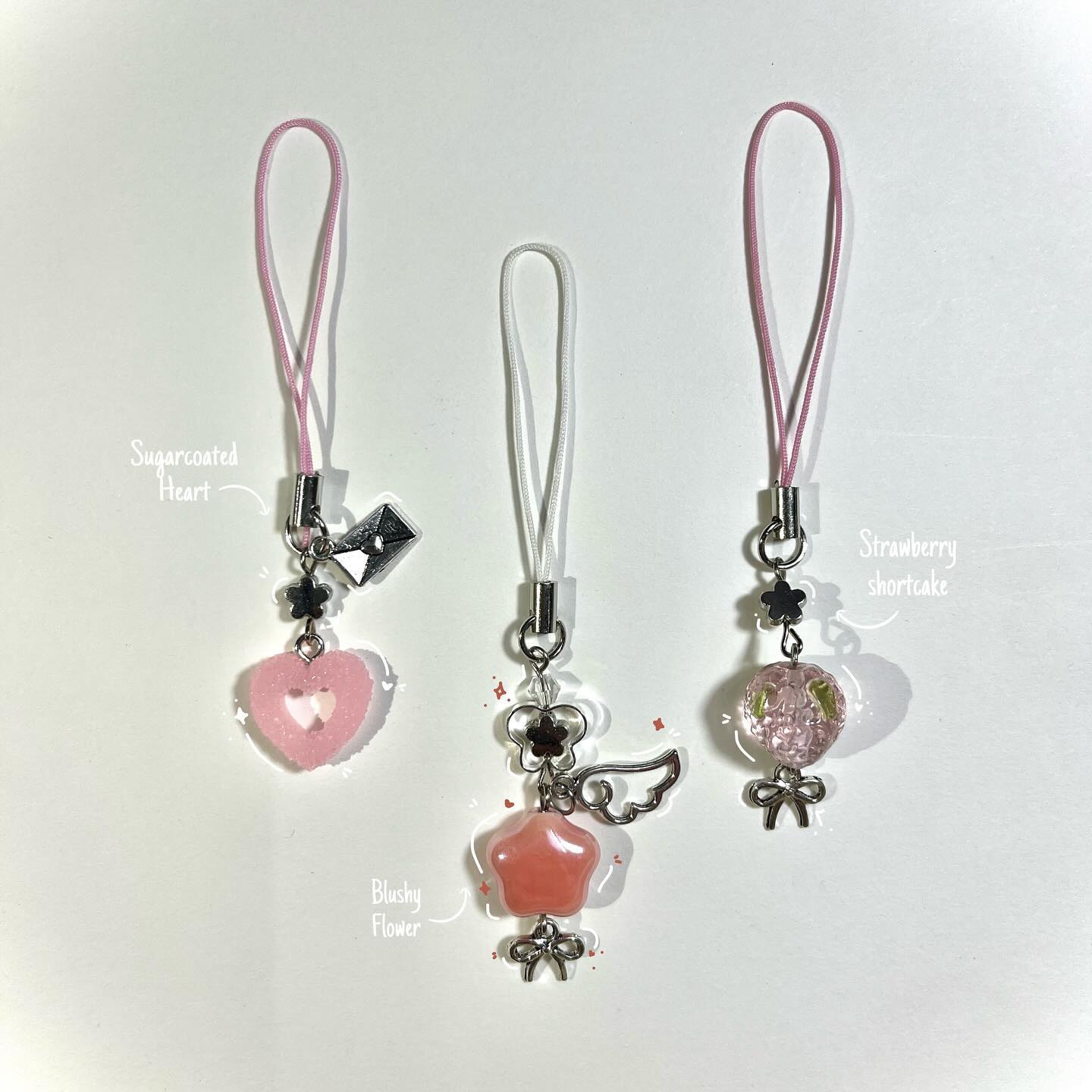 the image shows three cell phone charms on a white background. each charm has a different design and color scheme, but all have a cute, kawaii aesthetic. the charm on the left is a pink heart with a white outline, labeled "sugarcoated heart". the middle charm features a pink star with wings, labeled "blushy flower". the charm on the right is a clear bead with pink and green floral accents inside, labeled "strawberry shortcake". all charms have a strap for attaching to a cell phone or other device.