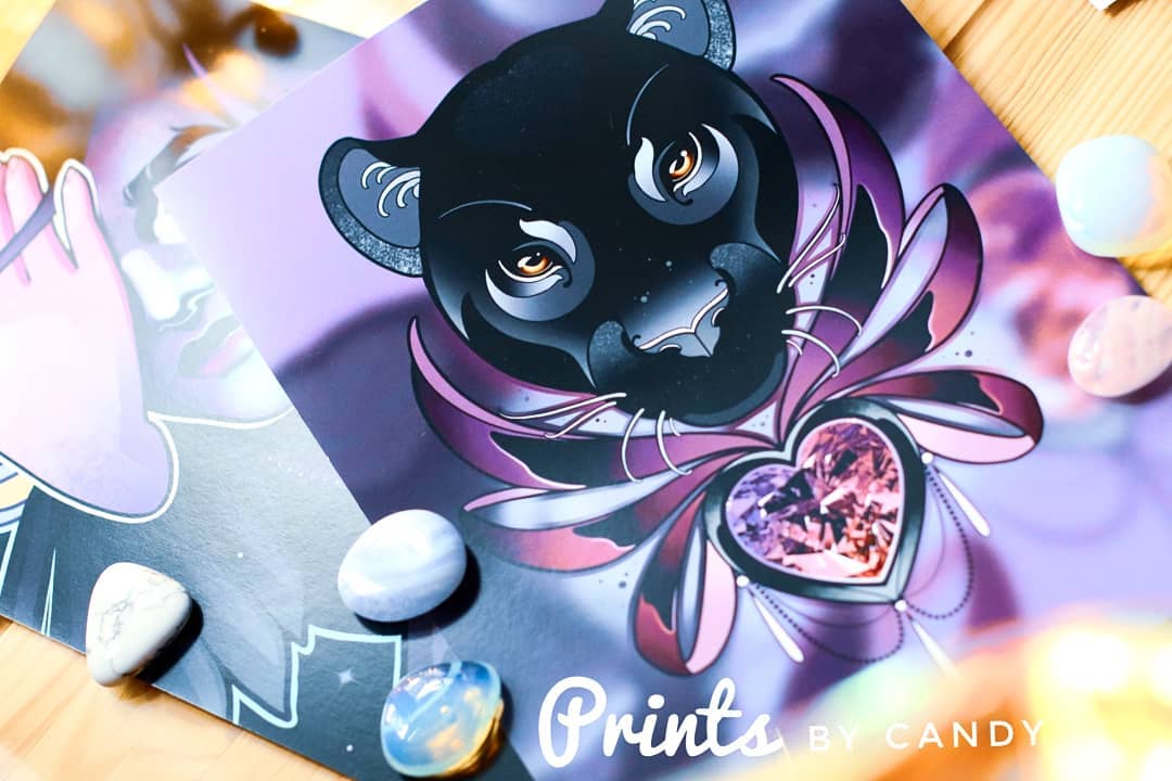 the image shows a colorful art print featuring a black panther head with golden eyes. the panther is adorned with pink wings and a heartshaped jewel in shades of pink and purple. the print rests on a wooden surface and is partially obscuring another print. there are several colorful stones scattered around the prints, adding to the mystical aesthetic.