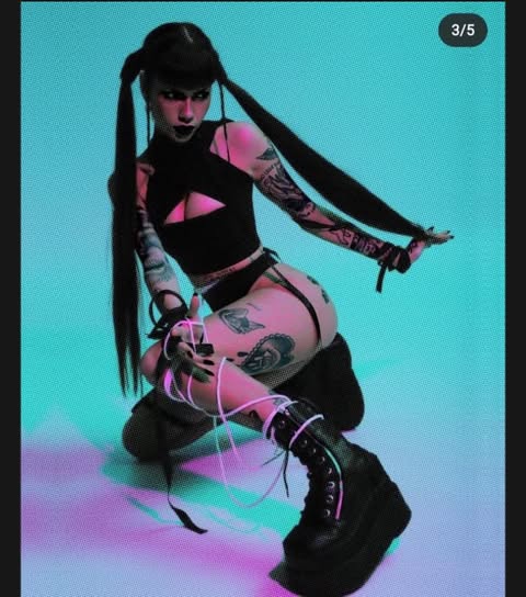 an edgy woman is kneeling in a photograph. she is wearing a black top with a cutout, highcut bottoms, and platform boots. she has pigtails, and she is covered in tattoos. strings are wrapped around her leg, leading up to her hand. the picture is under a blue and pink light. the image is marked "3/5" in the upper right corner.