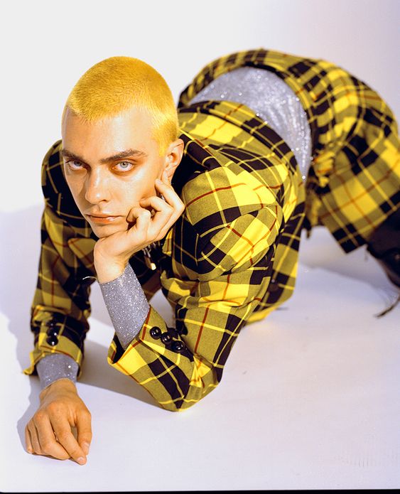 a male model with bleached yellow hair and striking blue eyes is posing on all fours in a studio setting. he is wearing a bright yellow and black plaid suit with sparkling silver cuffs. he is gazing intensely at the camera. the lighting is bright, highlighting the vivid colors of his outfit.