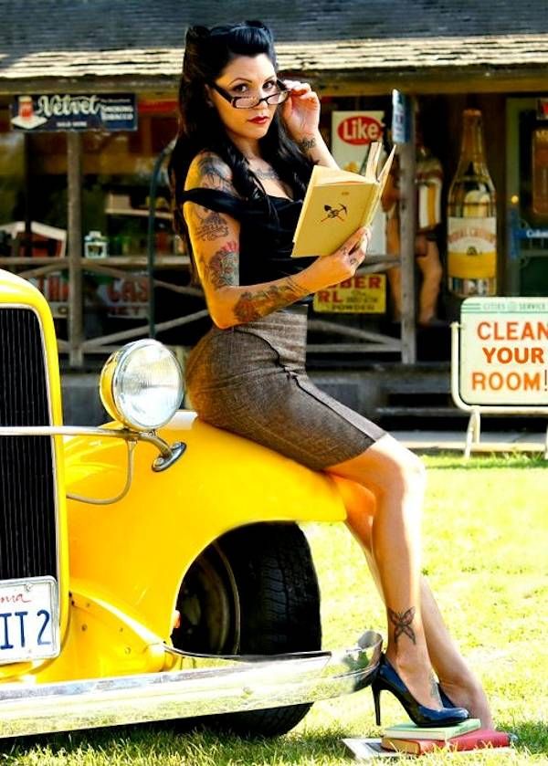 a pinup style woman with tattoos and victory rolls sits on the hood of a yellow vintage hot rod.  she's wearing a pencil skirt, high heels, and glasses, and is reading a book. the setting appears to be a vintage gas station or roadside diner, indicated by the signs and architecture in the background. the overall aesthetic is very retro and 1950s inspired.