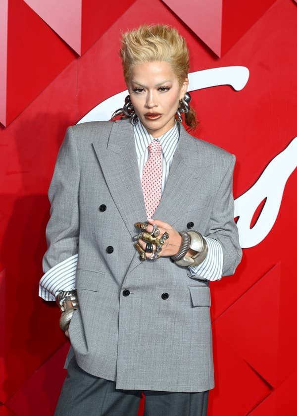 the image shows rita ora posing on a red carpet. she is wearing an oversized gray blazer with black buttons over a striped shirt and a pink tie. she is wearing multiple rings and bracelets on her hands and has her hair styled in a spiky fashion. the background is red with a white design.