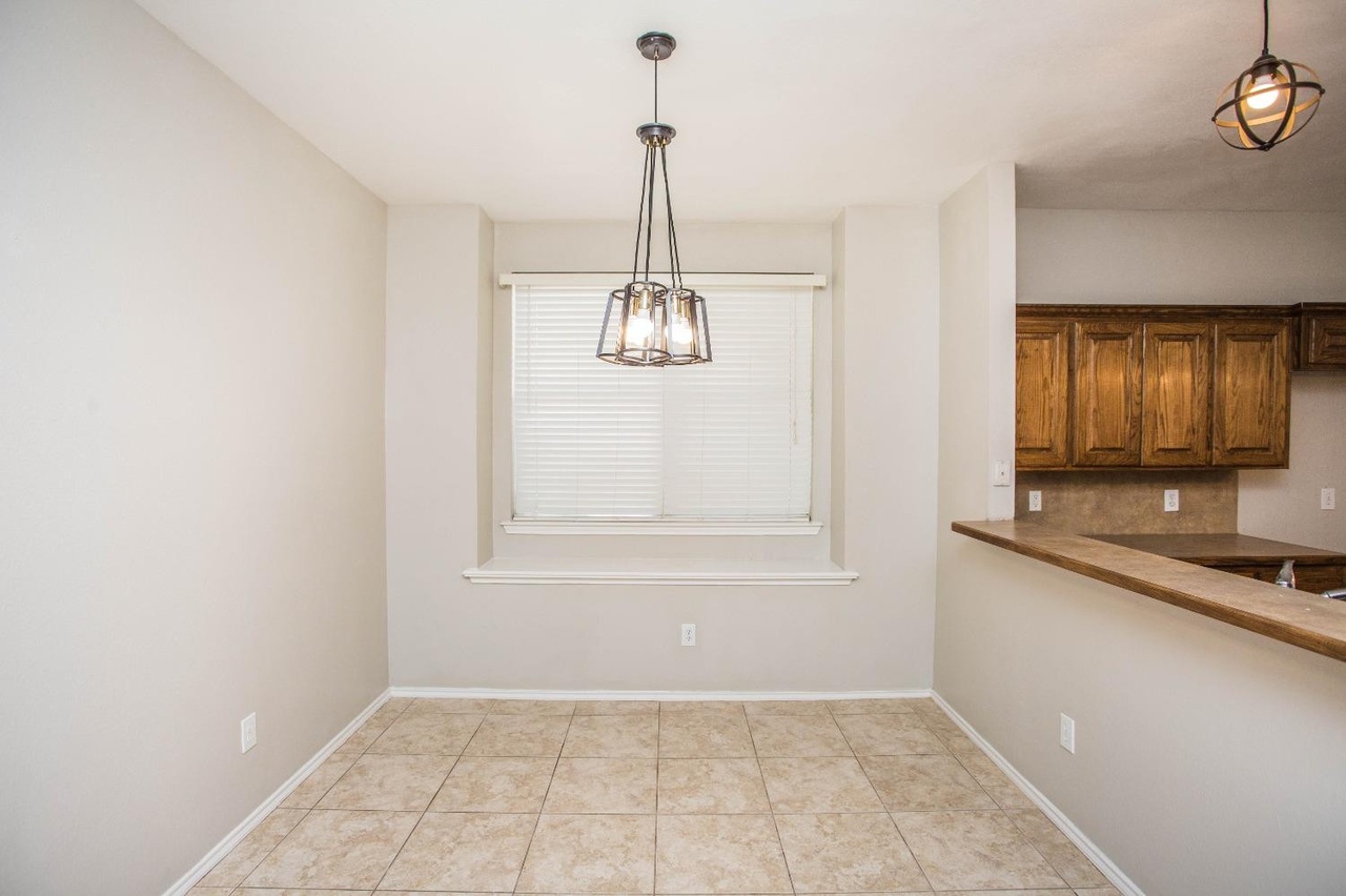 a welllit dining room with a window and chandelier, tiled flooring, and a view into a kitchen with wooden cabinets and a breakfast bar. the color scheme is neutral.