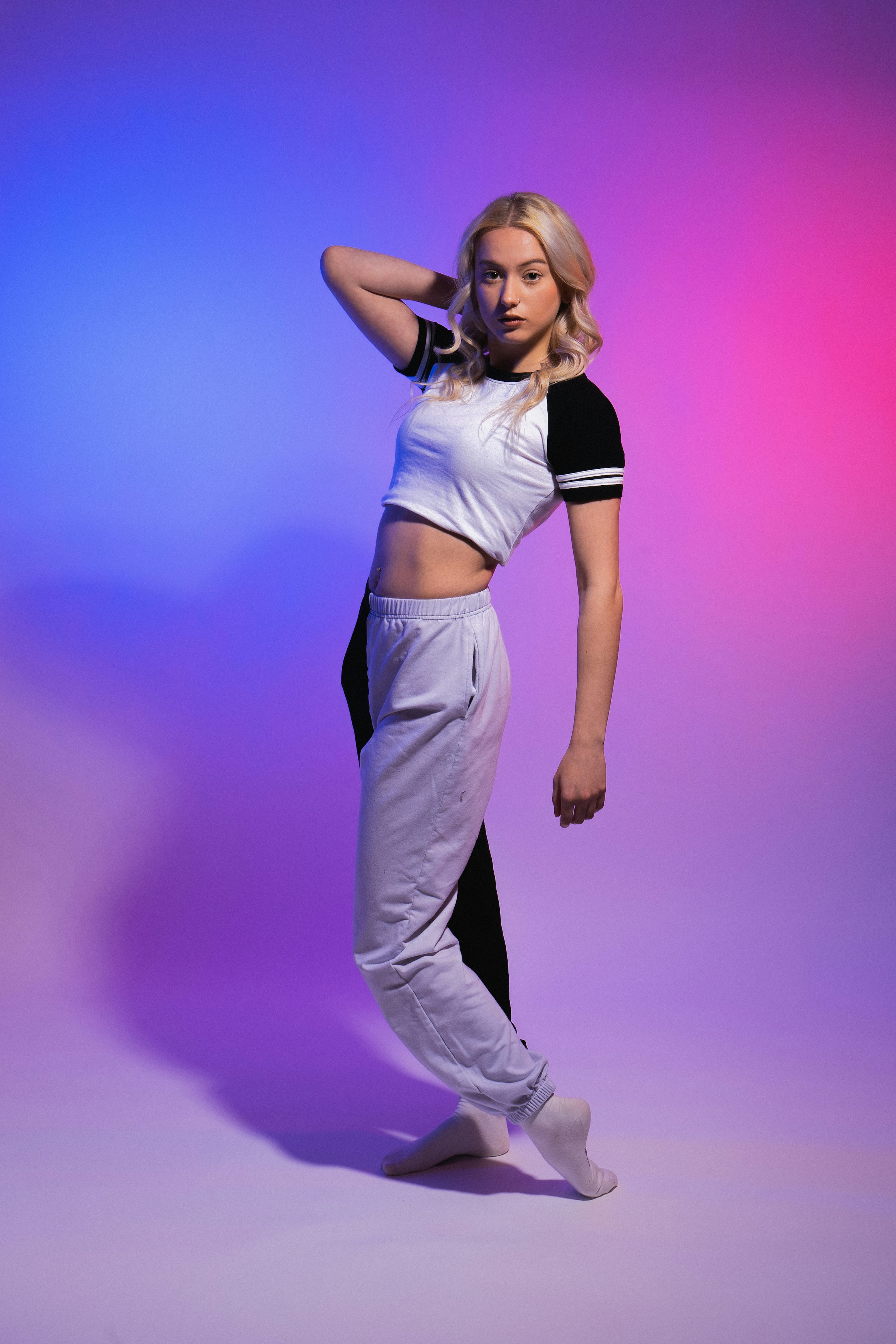 the image shows a young blonde woman in a stylish, casual outfit posing against a vibrant purple and blue gradient background. she is wearing a white and black cropped top and twotoned sweatpants (grey and black), which together create a modern sporty look.  her pose is relaxed yet confident, with her hand casually in her hair, suggesting a sense of ease and style.  the lighting is dramatic and neonlike, highlighting her features and outfit. the overall aesthetic is trendy and fashionforward.