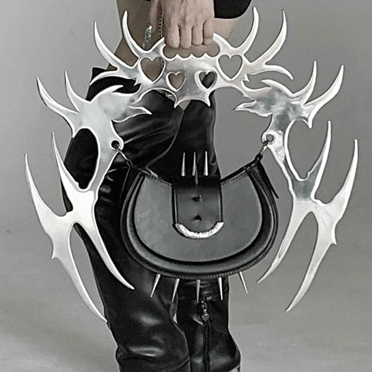 a person is holding a black handbag with silver spikes and a large, elaborate silver handle featuring heart shapes and spiky extensions. the person is wearing black leatherlike pants and a black top. the background is a plain gray.
