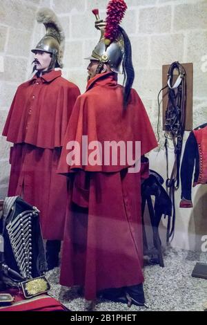two soldiers in traditional red military uniforms are standing in an indoor space that appears to be a museum or a historical setting. both are wearing  matching red coats and have distinctive hats and helmets. the soldier on the left is holding a weapon that is partially obscured by his coat. the soldier on the right has a sword on his hip and is standing at ease with his arms crossed.