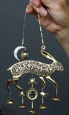 a person is holding a golden hare necklace. the necklace is ornate with cutouts on the body of the hare, and various trinkets hanging from it, including a frog, sun, and mouse. there is also a crescent moon hanging above the hare with a stone ball with a face. the necklace is hanging from a chain held in the person's hand. the background is a solid dark gray.