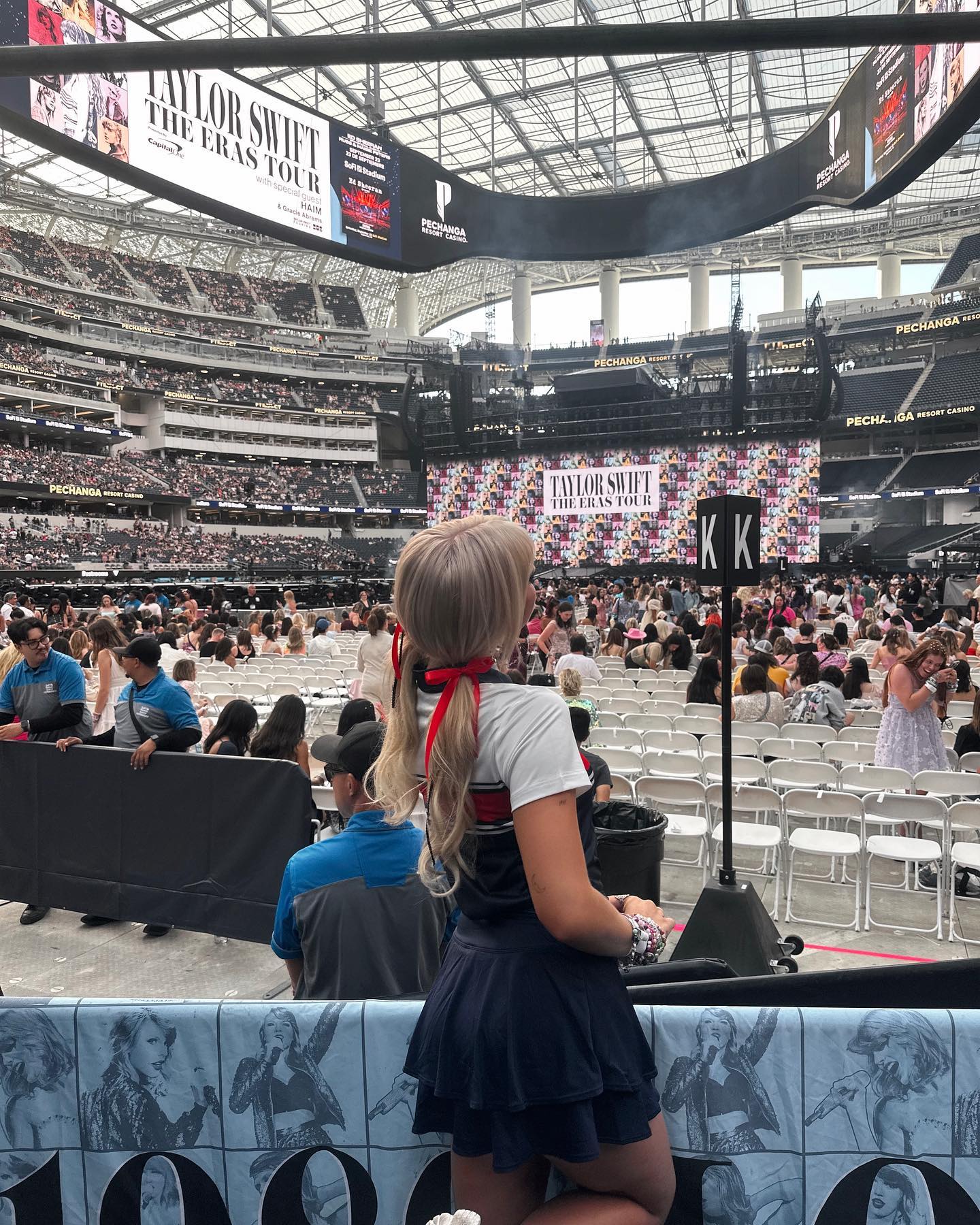 a fan in a cheerleader outfit stands in front of a banner at the taylor swift eras tour concert in a stadium full of people. the stage and large screens showing "taylor swift the eras tour" are visible in the background.