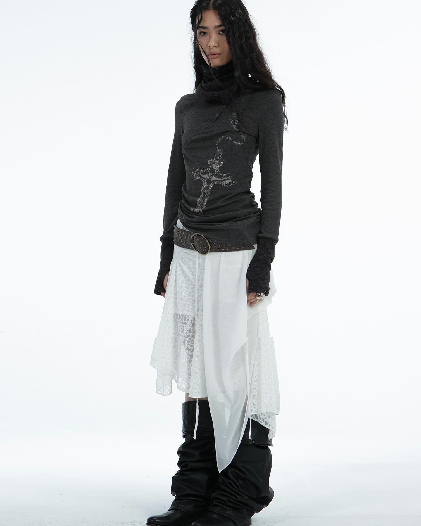 a fulllength studio shot of a woman modeling a dark grey longsleeved turtleneck shirt with a graphic design, a white layered skirt with lace detailing, a brown belt, and black high boots. the background is white.  the model is standing and posing.