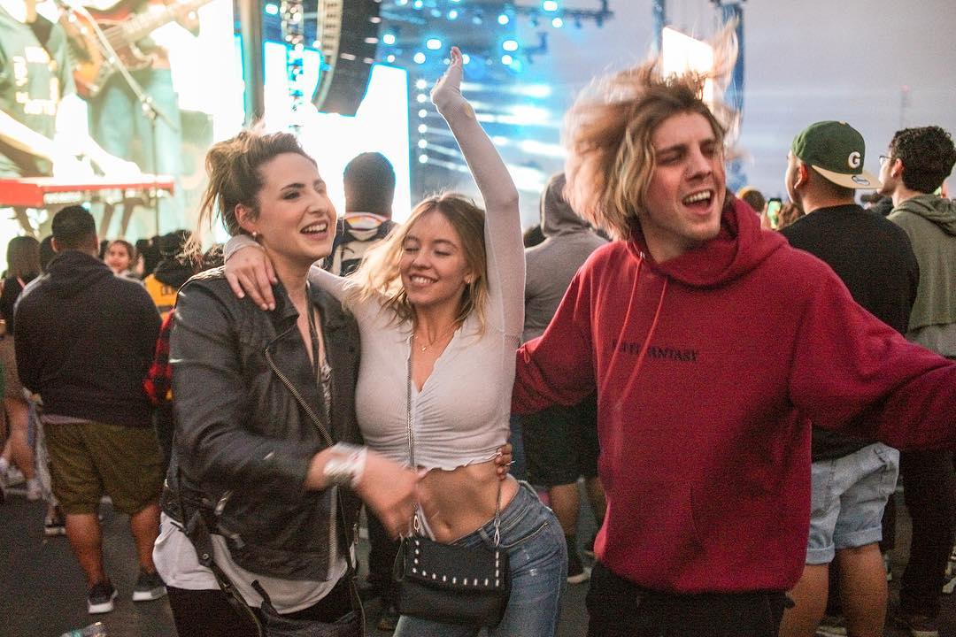 hree young adults, two women and a man, are joyfully dancing and interacting in a lively concert crowd. the women are embracing, one with her arm raised in excitement. the man in a red hoodie is also engaged in the celebration. the background is filled with a large audience at an outdoor music festival, under bright stage lighting.  the overall atmosphere conveys a sense of fun, energy, and shared enjoyment of a live music performance.