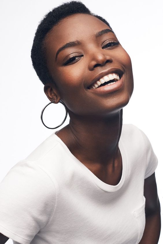 a young black woman with short curly black hair is smiling and looking at the camera. she is wearing a white tshirt and black hoop earrings. she has a beautiful complexion and is wearing no makeup. the background is white. she looks very confident and happy.