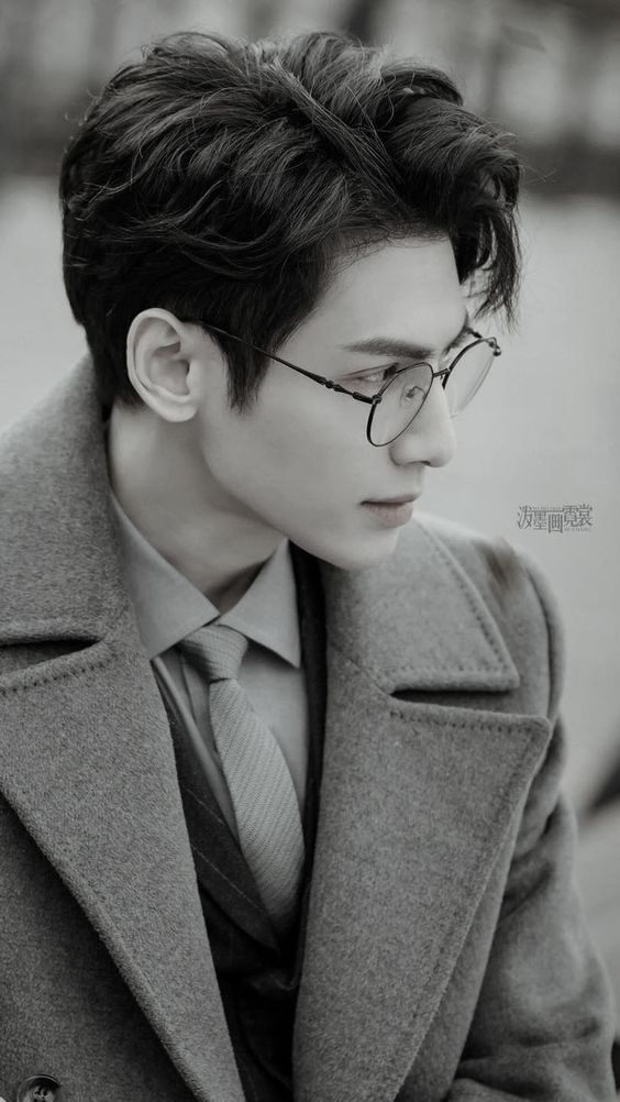 the image is a black and white portrait of a handsome young asian man wearing glasses and a suit. he is standing outside and looking to the side. the lighting is natural and the perspective is at eye level.