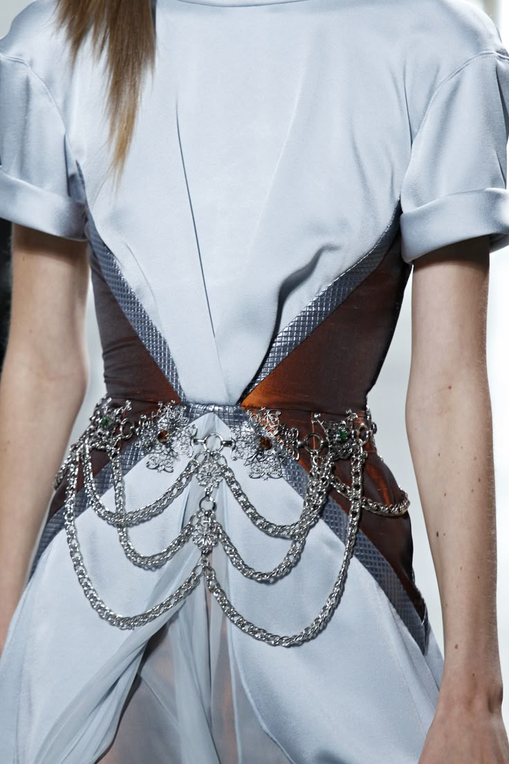 a closeup shot of a person wearing a light blue dress with short, rolledup sleeves and a brown, meshaccented bodice. a silver chain belt with ornamental details and dangling chains cinches the waist. the dress appears to be constructed with draping fabric in the front.