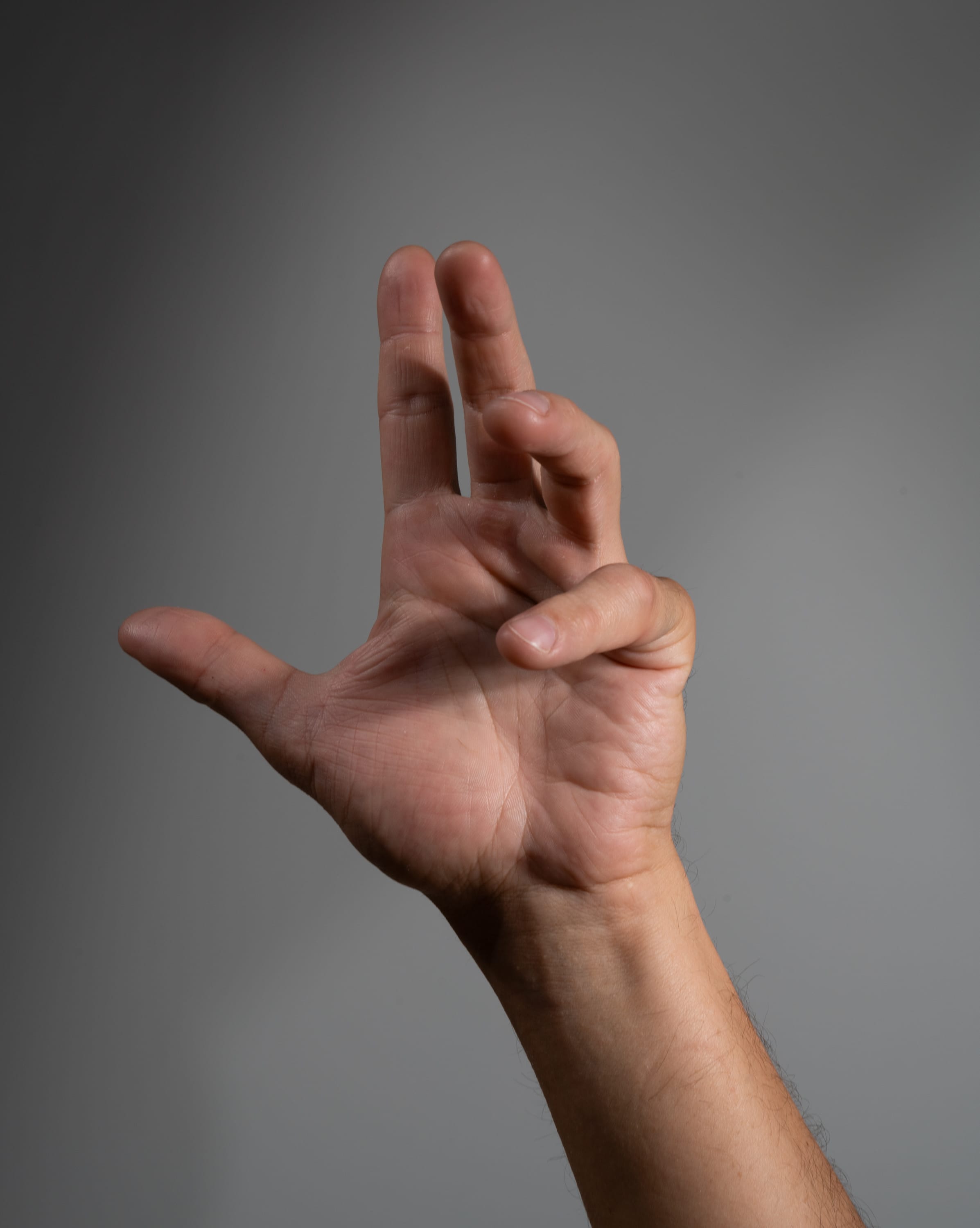 a closeup image of a man's hand making a gesture against a neutral gray background. the hand is extended with the index and middle fingers raised and slightly curled, while the ring finger and pinky are tucked into the palm. the light source appears to be natural, coming from the upper left, creating soft shadows on the hand and arm.