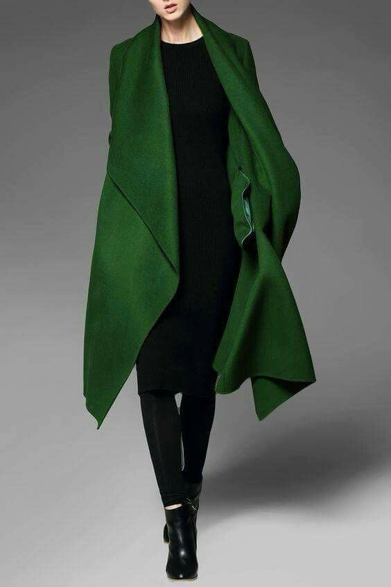 a woman in a long, green coat, with a black dress and black boots, standing in front of a gray background, looking at the camera. she is wearing a fashionable and elegant outfit.