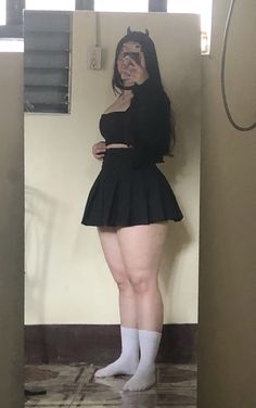 a young girl in a black pleated skirt and white socks  standing in front of a mirror. she is wearing a black top and  and is smiling and looking at her reflection.  the background is a mirror, and there is a white wall behind the mirror.