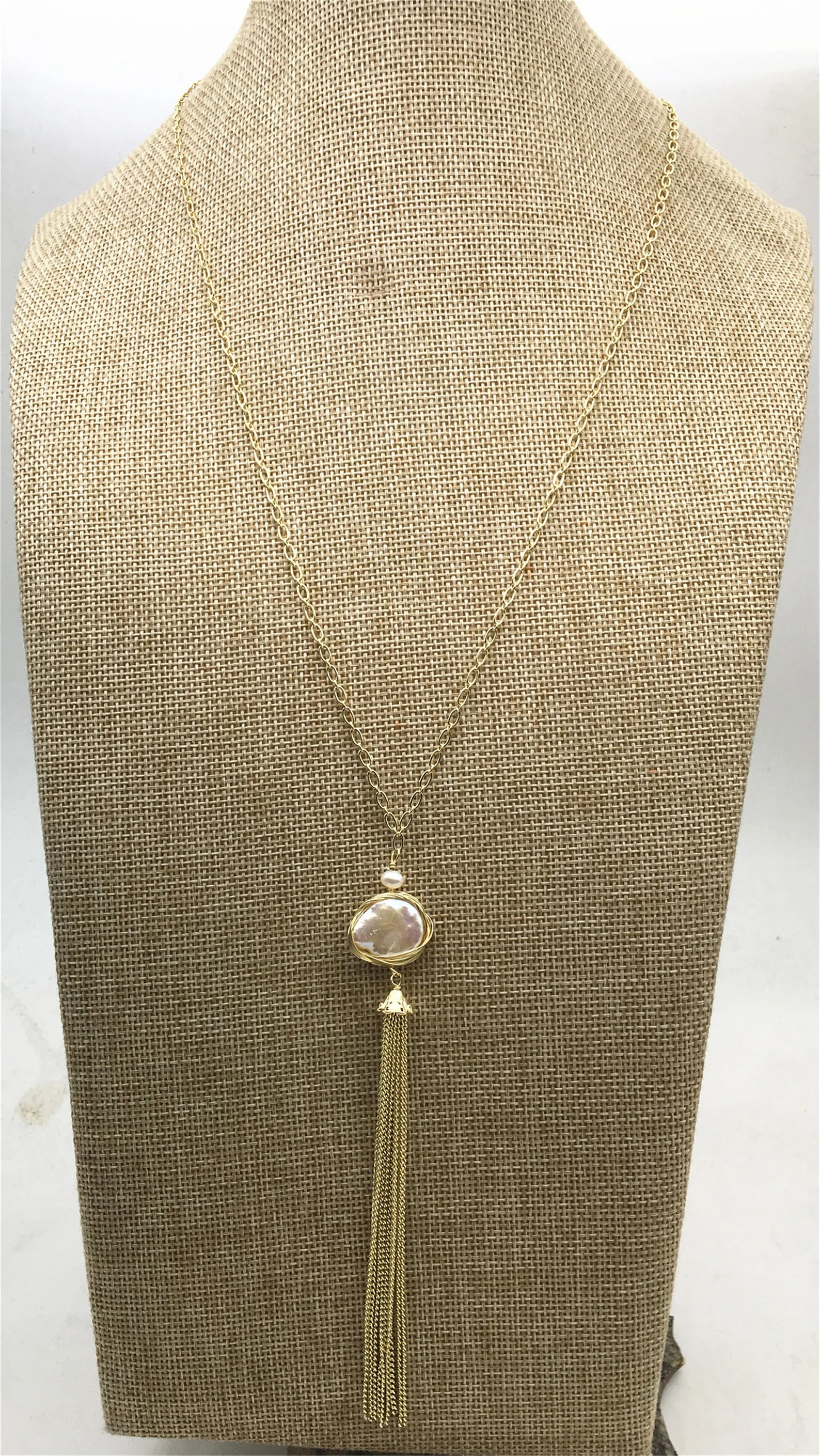 the image shows a gold chain necklace with a large round pearl pendant and a tassel. the necklace is displayed on a textured mannequin bust.content: necklace, jewelry, mannequin bust, fashion accessorypeople: environment: indoorlighting: artificialperspective: eyelevelkw: gold chain necklace, pearl pendant, tassel, fashion, accessory, jewelry, mannequin bust, elegant, stylish, feminine