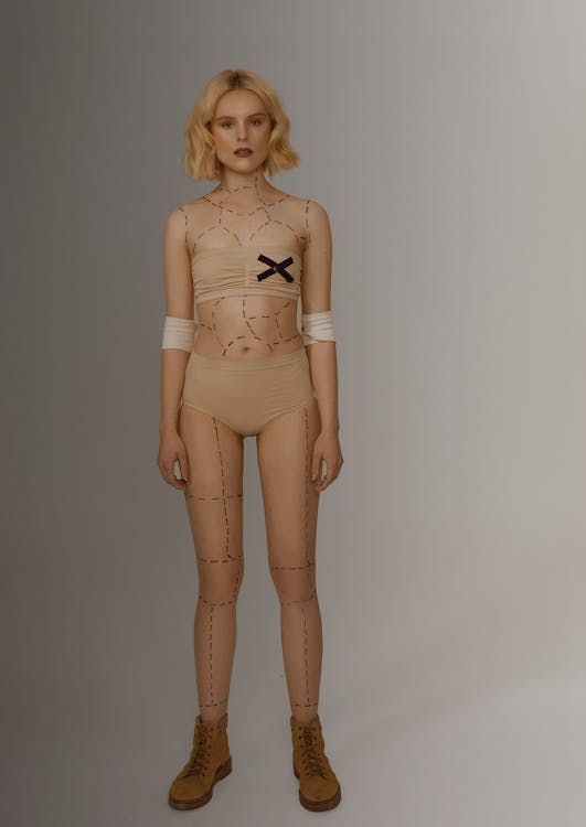 a woman stands facing forward in a studio shot, wearing tan underwear and a tan crop top. her body is marked with dotted lines resembling plastic surgery markings. she has a black "x" on her chest and bandages wrapped around her upper arms. she's wearing brown boots.