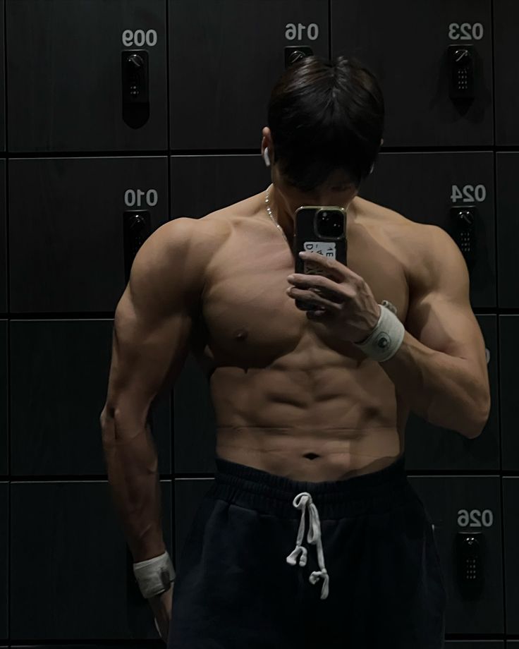 a muscular young asian man takes a shirtless selfie in a dark gym locker room. he's showing off his welldefined abs and sculpted physique. the background consists of darkcolored lockers with numbers on them.  he's wearing dark sweatpants and a wristband. the image has a moody, somewhat shadowy atmosphere.