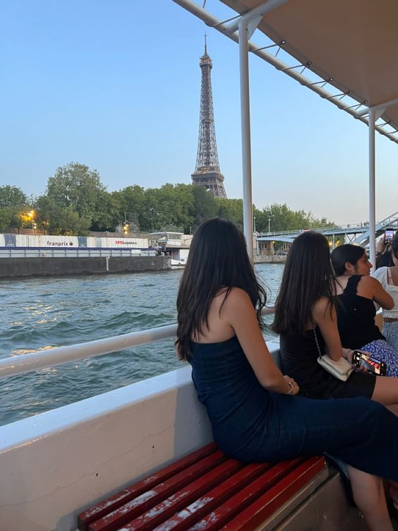 the image shows a view from a boat on the seine river in paris. in the background is the eiffel tower. in the foreground are three women sitting on a bench on the boat. the water is blue and the sky is clear. the lighting is natural daylight.