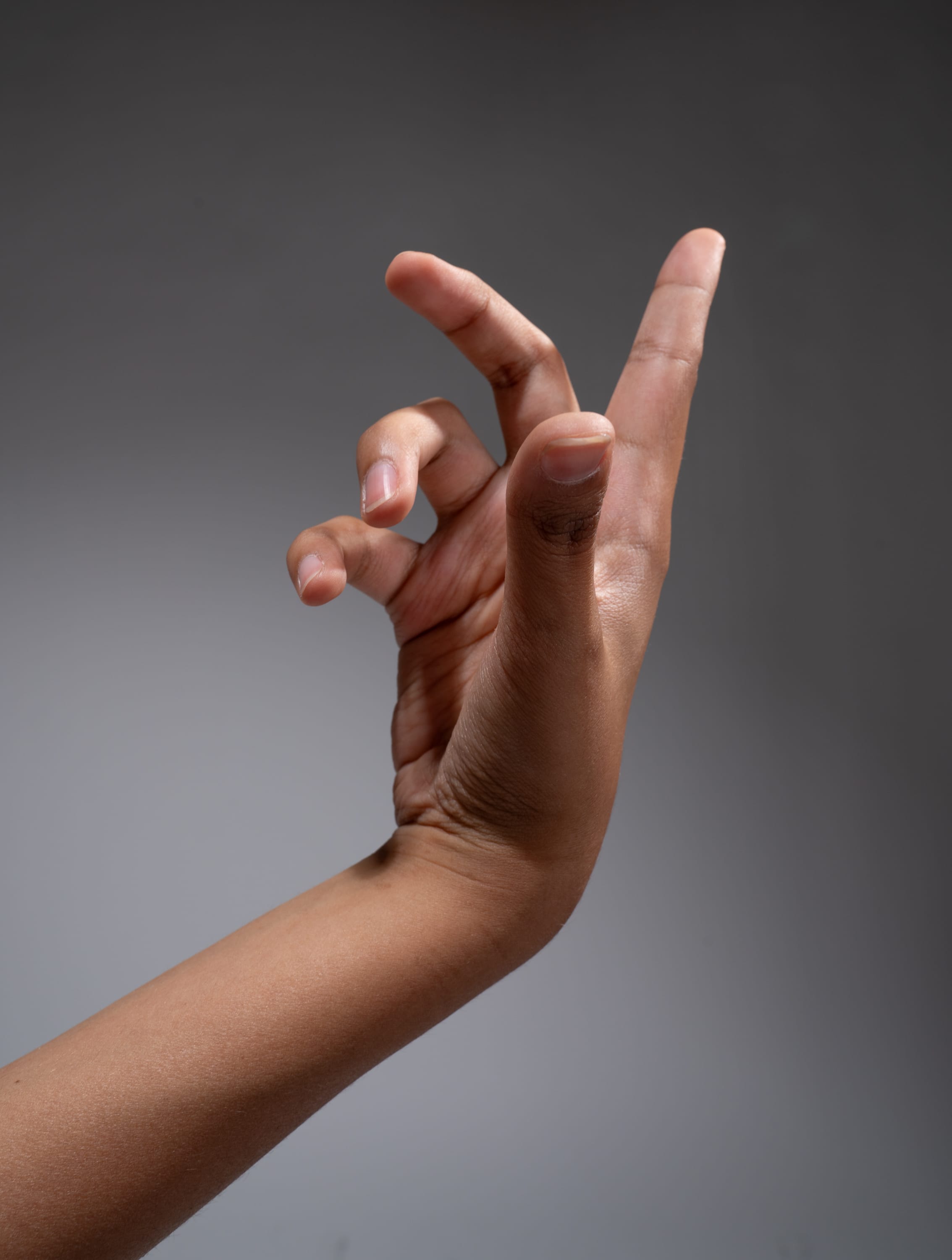 a moderately tan hand positioned against a gray backdrop is slightly raised and extends its fingers outward. the fingers are partially curled inward, creating a distinct clawlike hand gesture.