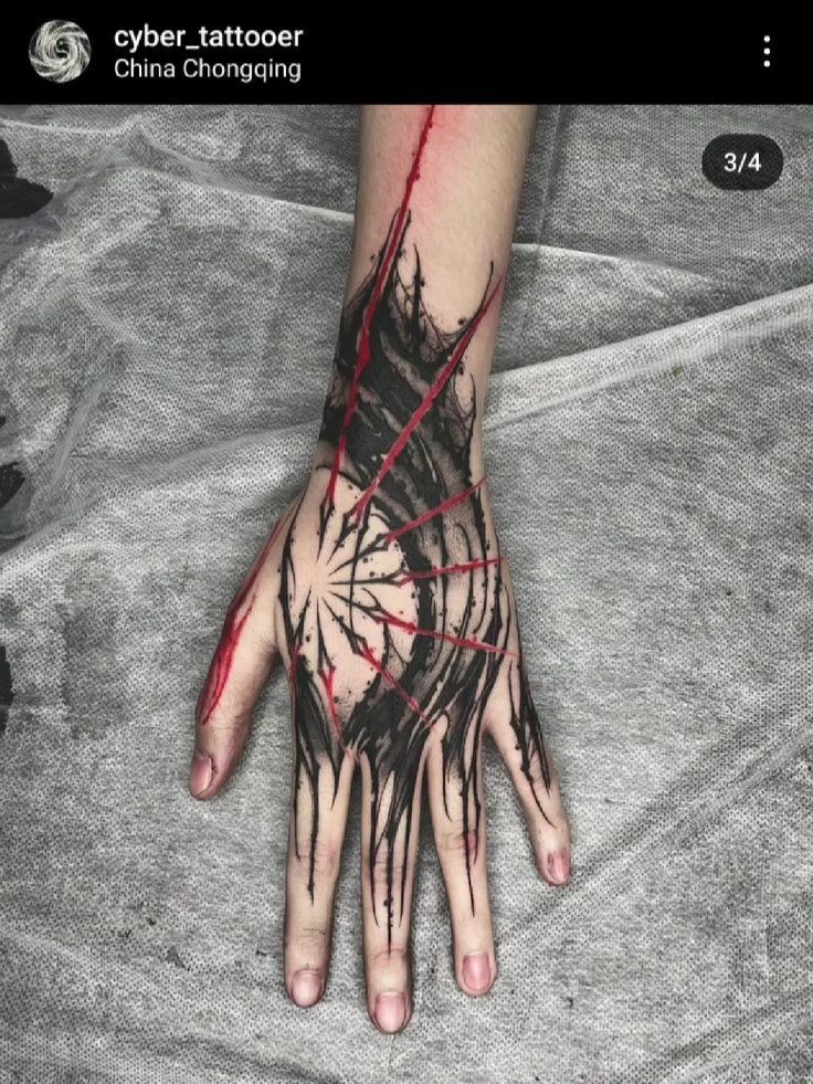 an arm and hand with a black and red abstract tattoo are shown against a grey textured background. the tattoo covers the wrist, the back of the hand, and extends onto the fingers. the tattoo is primarily black with red accents, giving the impression of slashes or cuts. the arm appears to be resting on the grey fabric. the upper left corner of the image has the name of the tattoo artist and location.