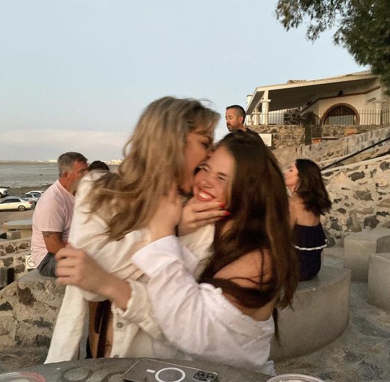 two young women are hugging and laughing together on a beach. they are both smiling and look very happy. one of the women is kissing the other woman on the cheek. they are both wearing white shirts and have long dark hair. the women are standing in front of a body of water with a sunset in the background. there are other people in the background.