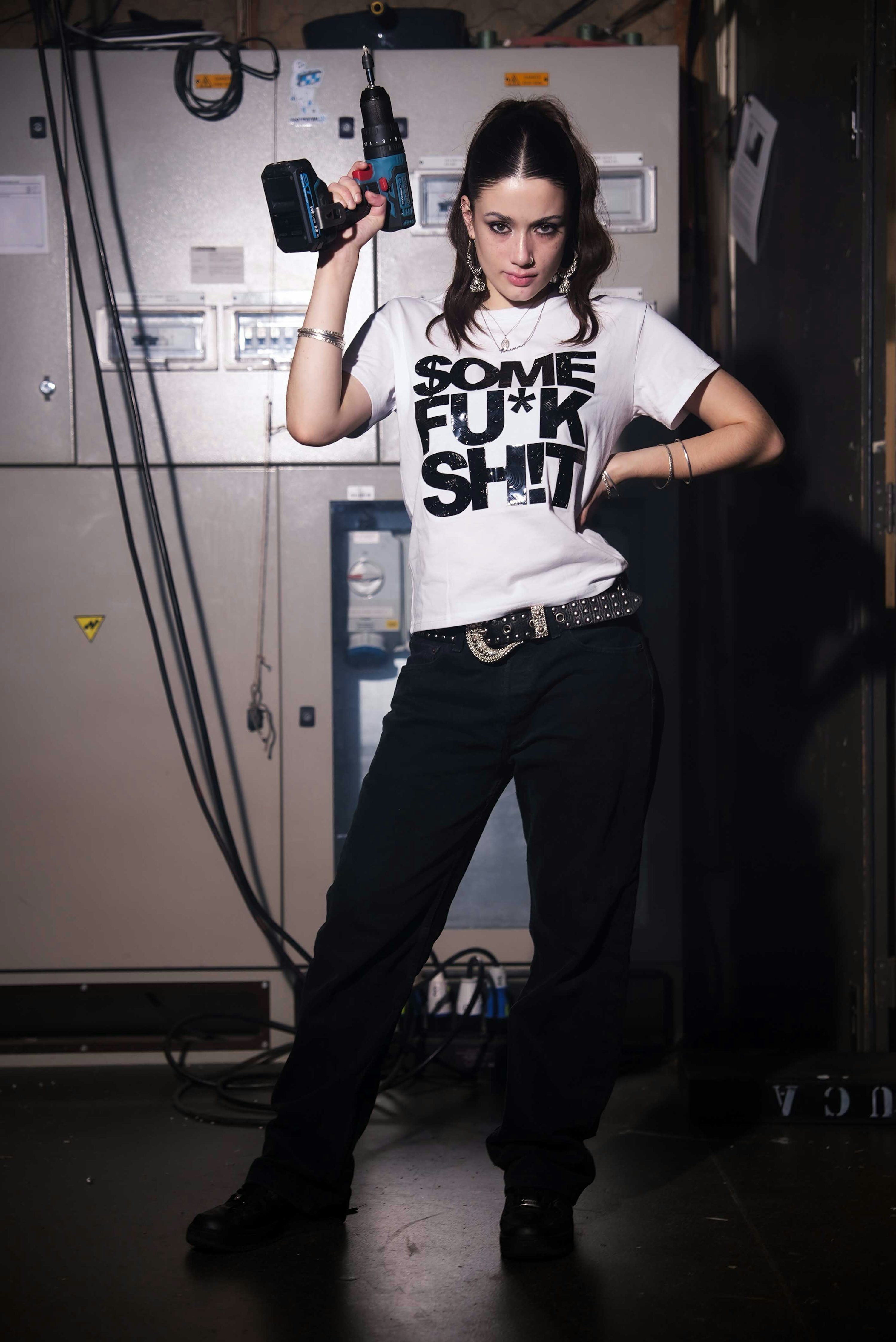 a woman is standing indoors, holding a drill and posing in front of an electricity box. she is wearing a white tshirt with bold black lettering that says "some fck sht," black pants, a studded belt, and black shoes. she has a ponytail and is wearing jewelry. the background shows an industrial setting with an electricity box and wires. the room is dimly lit.