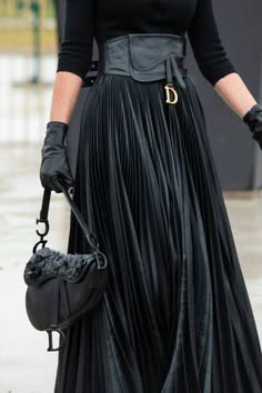 a woman is wearing a stylish black pleated skirt with a black leather belt and black leather gloves. she is also carrying a black handbag and a black jacket. she is standing on a street and looking at the camera. she is wearing a classic elegant outfit that is perfect for a night out or a special occasion.