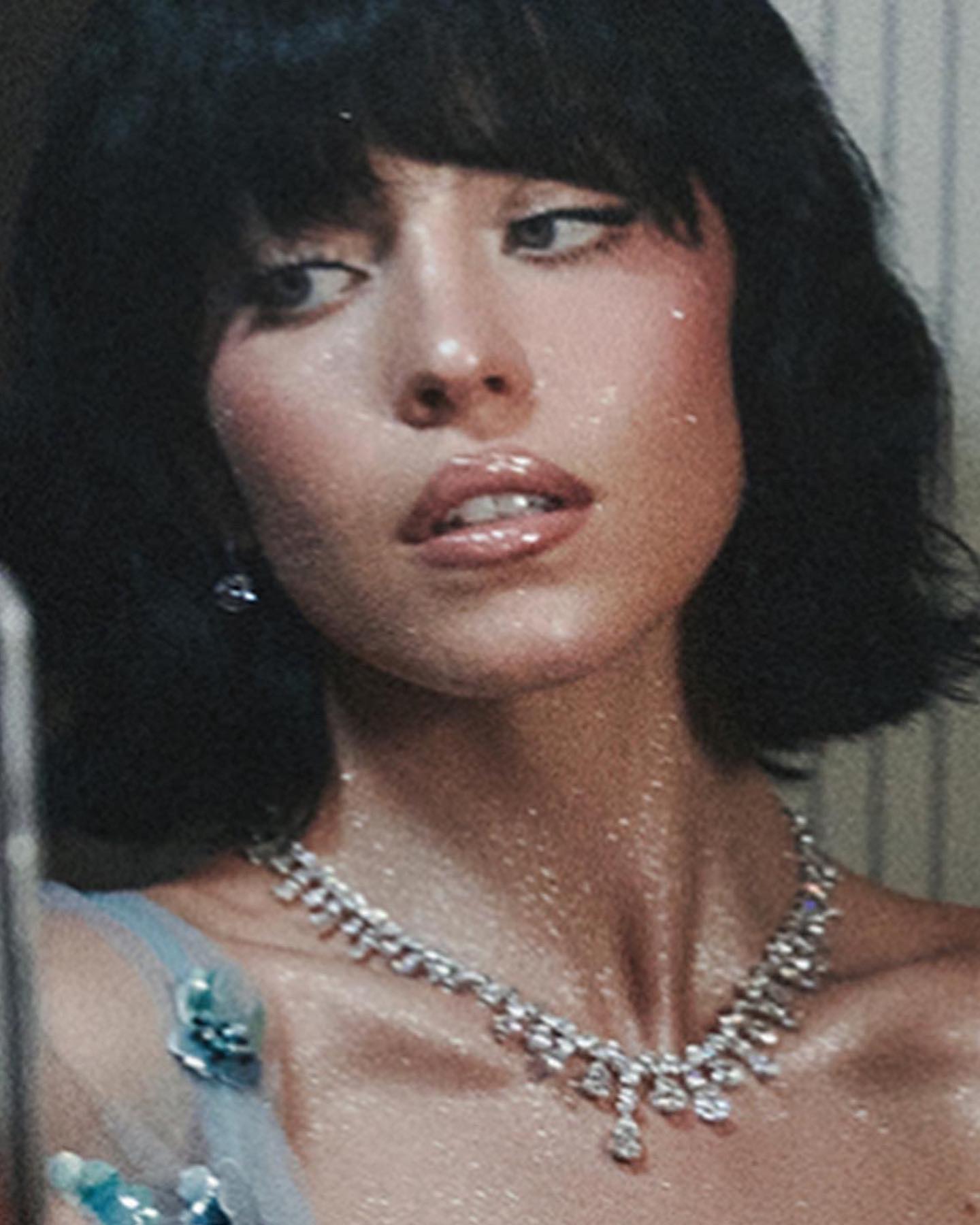 loseup portrait of a woman with dark bobbed hair and bangs. she is wearing a sparkly, light blue dress with a diamond necklace and has glitter on her skin. her makeup is noticeable, and she has a glamorous expression. the background is blurred but shows a hint of wallpaper. the overall style is reminiscent of retro or vintage fashion photography.