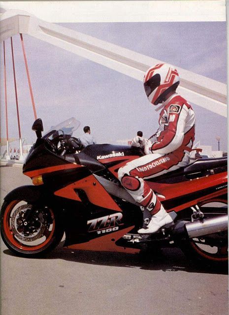 a kawasaki 77r 1100 motorbike is parked on a racetrack. the biker is wearing a white and red racing suit and helmet. the bike is in the foreground and the biker is sitting on the bike with his left leg on the ground. the bike is red, black and white, and has the kawasaki logo on it. the rider is looking to the left side of the image.  the background is blurred.