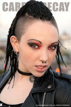 a woman with a shaved head and dreadlocks poses in a leather jacket. she has many piercings, including nose and lip rings, as well as multiple ear piercings. her eye makeup includes red eyeshadow. the text "carb decay" is partially visible at the top of the image.