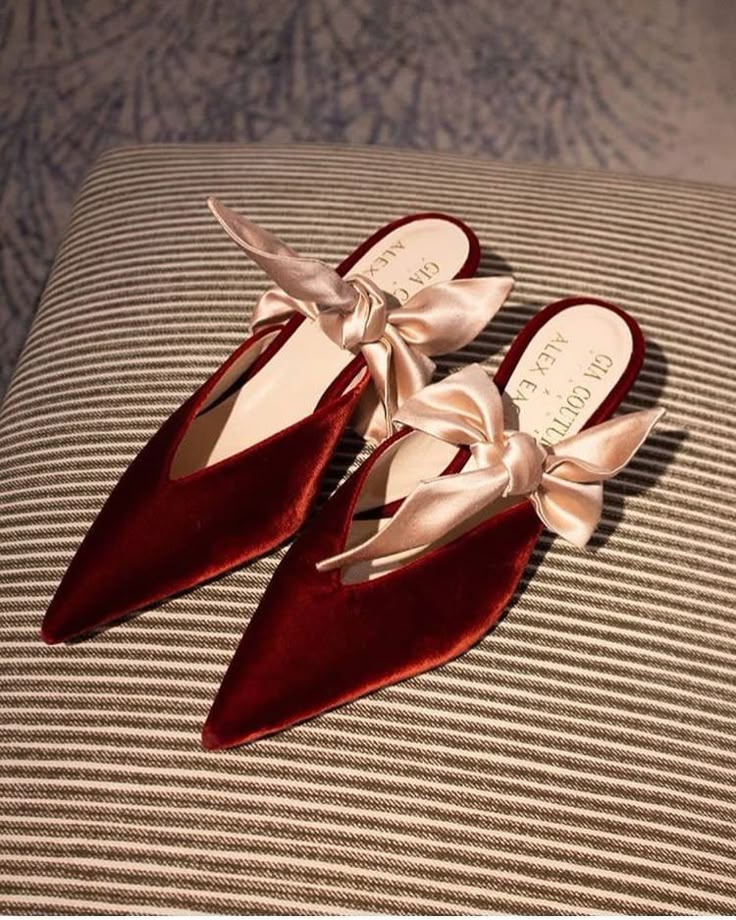 two maroon velvet pointedtoe mules with a light beige bow on each shoe are displayed on a striped cushion. the shoes are positioned at an angle, showcasing their design and the elegant bow detail. the stripes on the cushion provide a contrasting background to the rich color and soft texture of the shoes. the words "gia couture" and "alex eagle" are partially visible on the insole of each shoe.