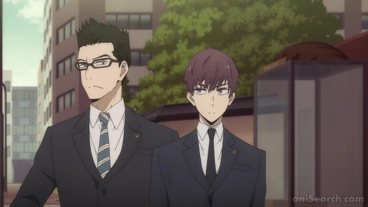 the image shows two men in suits standing on a city street. the man on the left is taller and has short black hair and glasses. he is wearing a black suit with a white shirt and a blue tie. the man on the right is shorter and has short brown hair. he is wearing a black suit with a white shirt and a black tie. both men have serious expressions on their faces. the background of the image is a city street with buildings and trees.