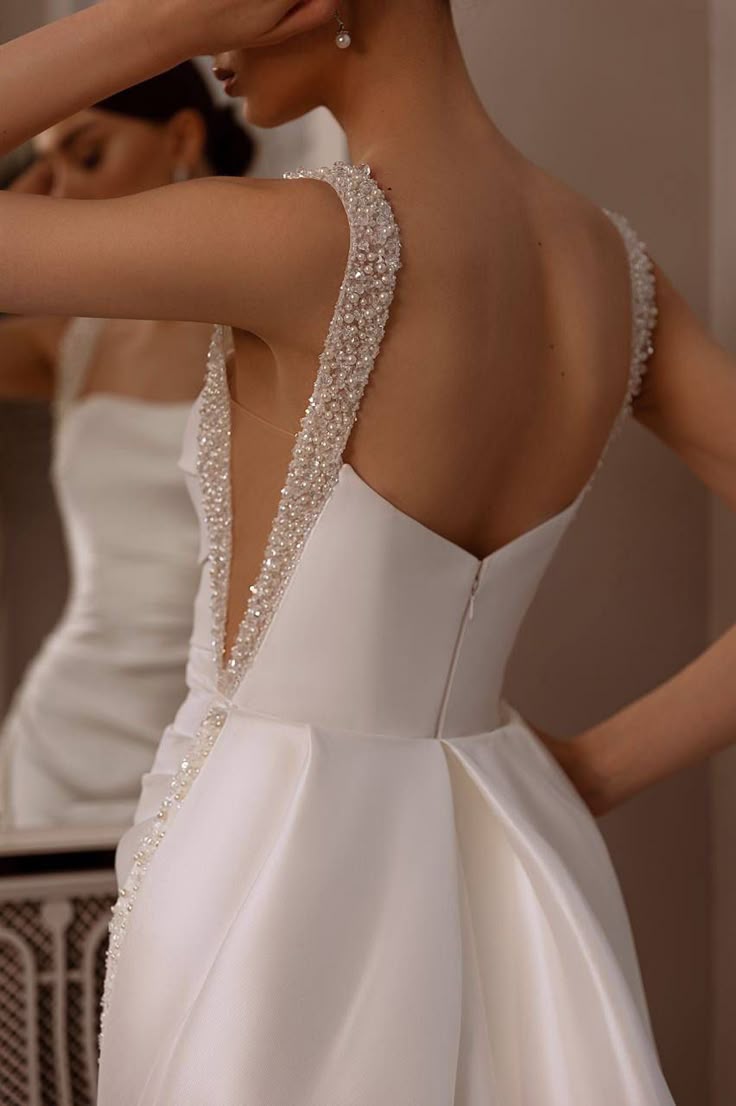 a woman is wearing a white satin wedding dress with beaded straps and a low back. the dress has a zipper in the back. a woman is partially visible in the mirror. the woman is partially covering her face with her hand. she is wearing pearl earrings.
