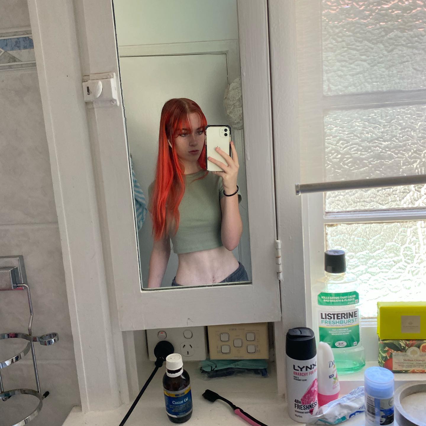 a young woman with brightly dyed red hair, styled with a parted fringe, takes a mirror selfie in her bathroom. she's wearing a green crop top and low waisted pants, revealing her slim figure. she holds her phone casually in front of her face, partially obscuring it from view. the bathroom is brightly lit by natural light streaming in through a window reflected in the mirror behind her. on the counter in the foreground, various toiletries and hygiene products are visible, including mouthwash, deodorant, and a bottle of castor oil. the overall impression is one of casual, everyday life.