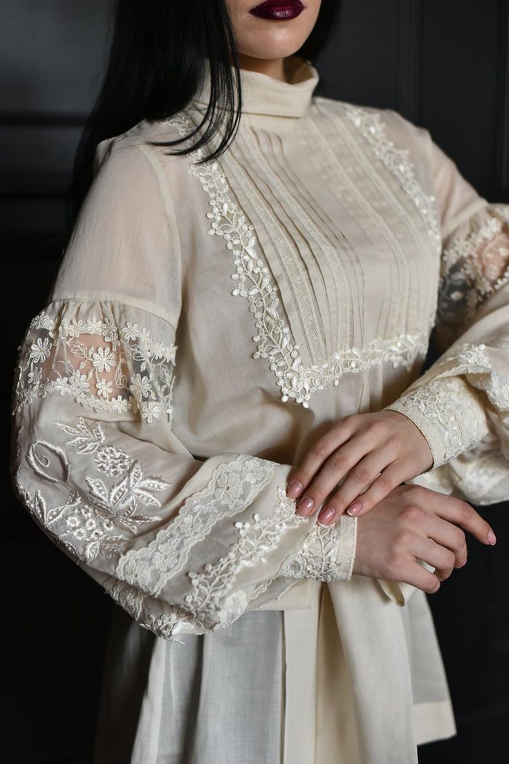 loseup view of a woman wearing an elegant, vintagestyle creamcolored blouse. the blouse features intricate lace detailing along the cuffs, collar, and placket.  the fabric appears sheer and delicate. the woman has dark hair and is wearing dark burgundy lipstick. her hands are clasped together, highlighting the delicate lacework on the sleeves. the background is dark and moody, allowing the clothing to stand out. the overall impression is one of refined elegance and classic style.