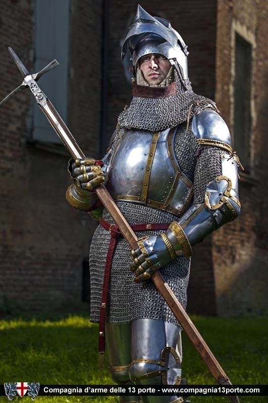 a knight in full plate armor, including a helmet with a pointed top, chainmail around the neck and shoulders, and polished metal armor on the chest, arms, and legs. the knight is holding a crossbow. the background includes a brick building and grass. the image also contains a watermark from "compagnia d'arme delle 13 porte."