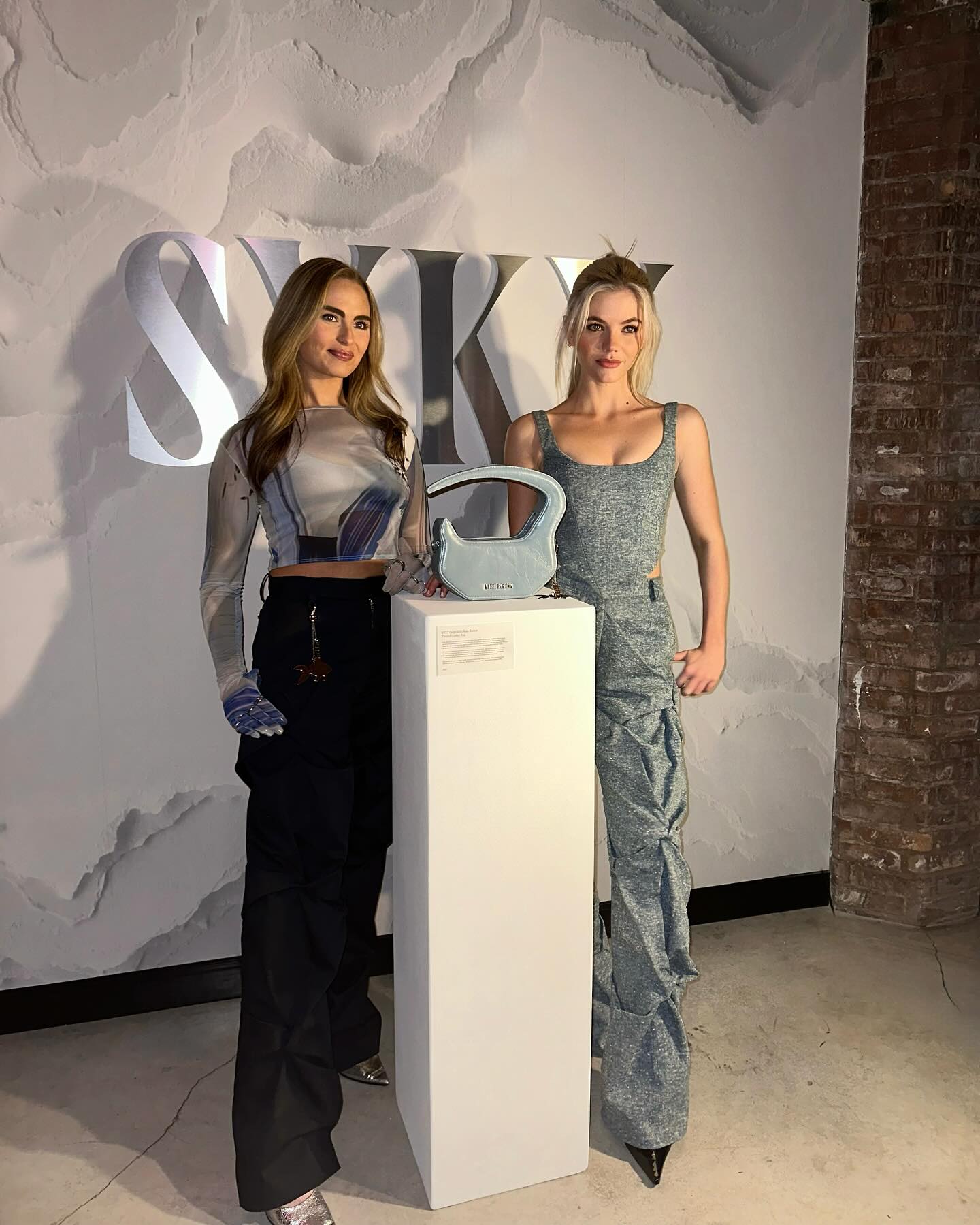 two women stand in an indoor studio setting beside a white pillar that is displaying a pale blue handbag. the woman on the left is wearing a long sleeve shirt with a blue design and black pants, and the woman on the right is wearing a gray crop top and pants set.