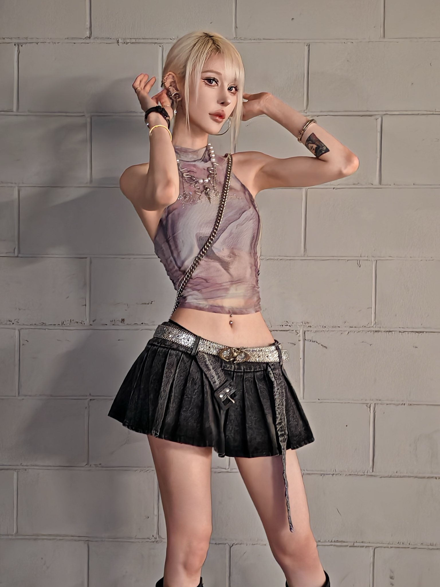 a young woman with short blonde hair and bangs is posing against a white brick wall. she is wearing a sheer, mauvecolored halter top, a black pleated denim mini skirt, a silver studded belt, several bracelets, earrings, and a necklace.  she is looking at the camera and has her hands near her head. she is wearing minimal makeup and has tattoos on her arms. her pose is stylish and confident. the overall image is a fashion portrait.