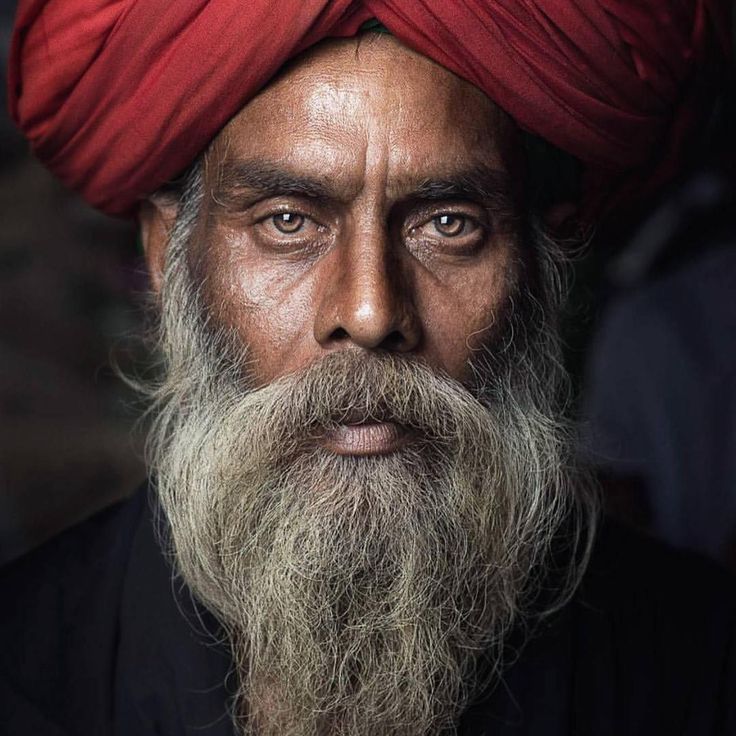 a closeup portrait of an older man with a weathered face, a long, gray beard, and a red turban on his head. he gazes directly at the viewer with intense, light brown eyes. his face is deeply lined and textured, suggesting a life of experience. the lighting highlights the details of his skin and beard, and he is dressed in dark clothing. the overall impression is one of wisdom, age, and cultural heritage.