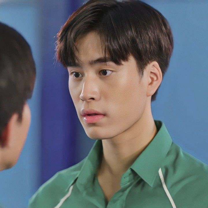 a young  asian man with short, dark hair and brown eyes wearing a green collared shirt is looking slightly to the left of the camera with a surprised expression.  he appears to be indoors. the background is out of focus and blurred, but appears to be mostly blue and white.