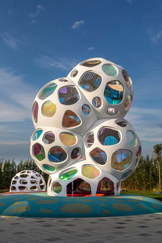 the image shows a modern, whimsical playground structure resembling a cluster of interconnected white bubbles with colorful, translucent windows. the structure stands out against a backdrop of a clear blue sky dotted with a few white clouds. set on a teal and blue rubberized ground, the playground is surrounded by a few trees, suggesting a park or outdoor setting. the image captures the structure from an eyelevel perspective, highlighting its organic shape and playful design.