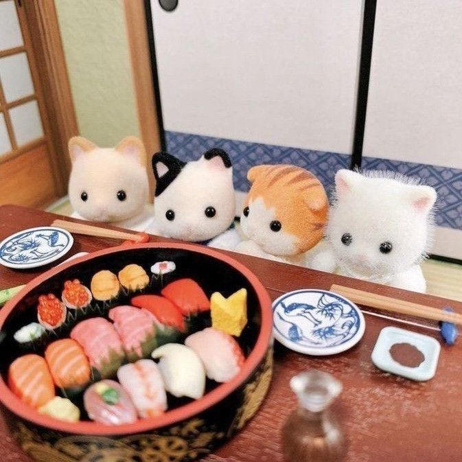 four calico critters cat figures are seated at a table with a platter of miniature sushi. the sushi includes various types of nigiri, such as salmon, tuna, roe, and tamagoyaki. there are also small dishes of wasabi and soy sauce. a pair of chopsticks rest on the table. two small plates with blue designs are also present.