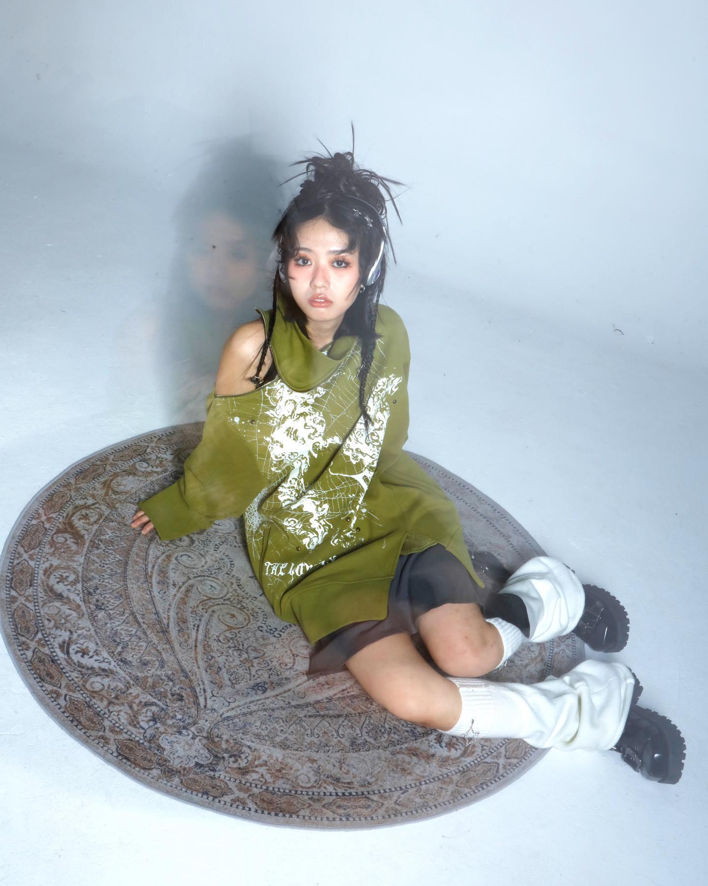 he image shows a young woman, likely a fashion model, posing for a photoshoot in a studio setting. she is seated on a large, round, patterned rug.  she is wearing an oversized, olive green sweater with a graphic print, styled with a partially offtheshoulder look.  the sweater is loosefitting and baggy.  she is also wearing chunky black boots and high white socks, adding to the grungeinspired look. her hair is styled in a messy bun.  the overall style is edgy and alternative. the background is a plain, light grey, allowing the model and her outfit to be the focal point of the image. the lighting creates a slightly moody atmosphere, while maintaining good visibility.