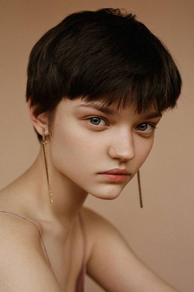 closeup portrait of a young woman with short, dark brown hair styled in a pixie cut. she's wearing long, delicate gold earrings and a simple mauvecolored strap top.  her expression is serious and her makeup is minimal, enhancing her natural features. the background is a plain, neutral beige. the overall aesthetic is elegant, minimalist, and sophisticated.