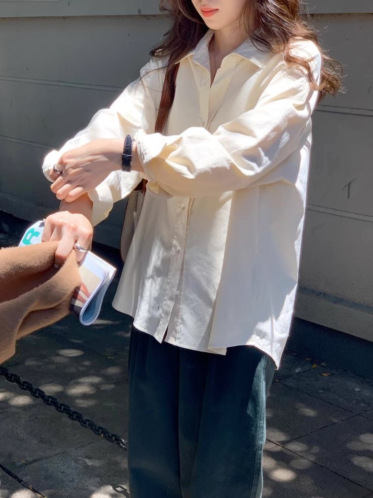 a stylish woman is posing outdoors in a creamcolored oversized buttondown shirt and dark green pants. the sun is shining on her, and she is holding a small notebook or book. her style is casual, minimalist, and chic. the photo appears to be taken in an urban setting, possibly a city street. the overall aesthetic is stylish and relaxed.